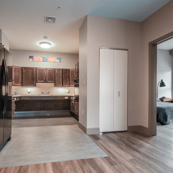 Open-plan accessible apartment with dark wood kitchen, stainless appliances, and view into bedroom with blue bedding.