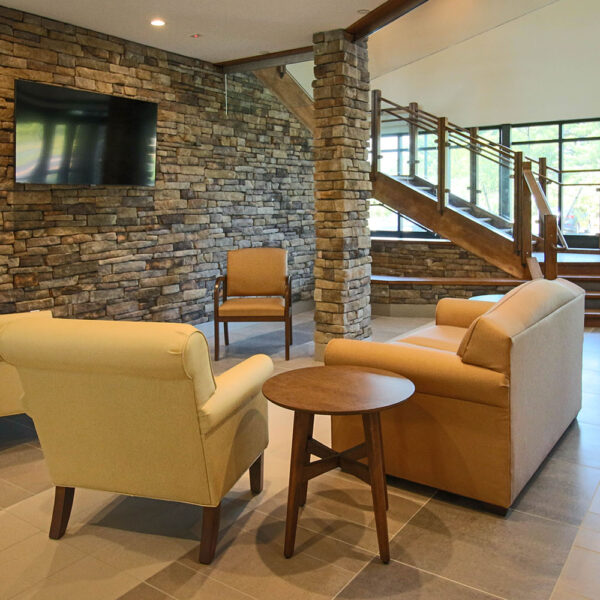 Chairs facing TV on stone covered wall in Shirley Meadows senior apartment building lobby with staircase and windows in background
