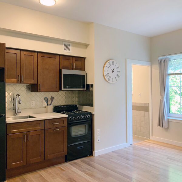 Compact kitchen with wooden cabinets, black appliances, hardwood flooring, and a view into an adjacent room.