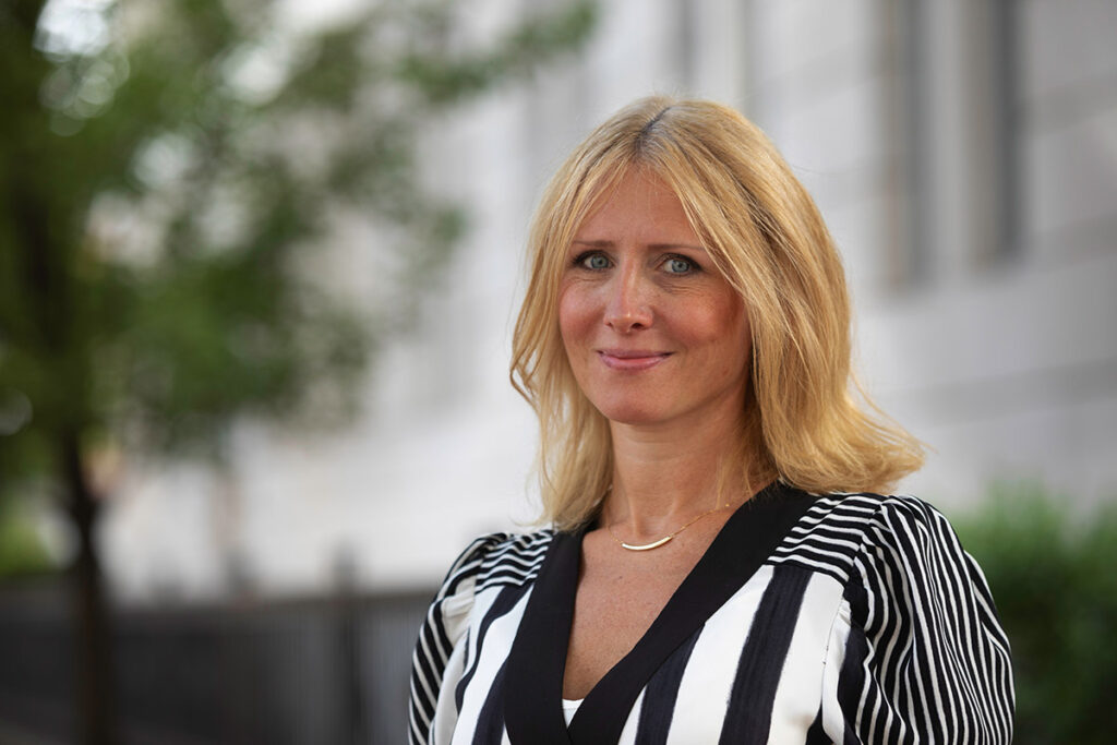 Person with blonde hair in a striped shirt, standing outdoors near trees and a building.