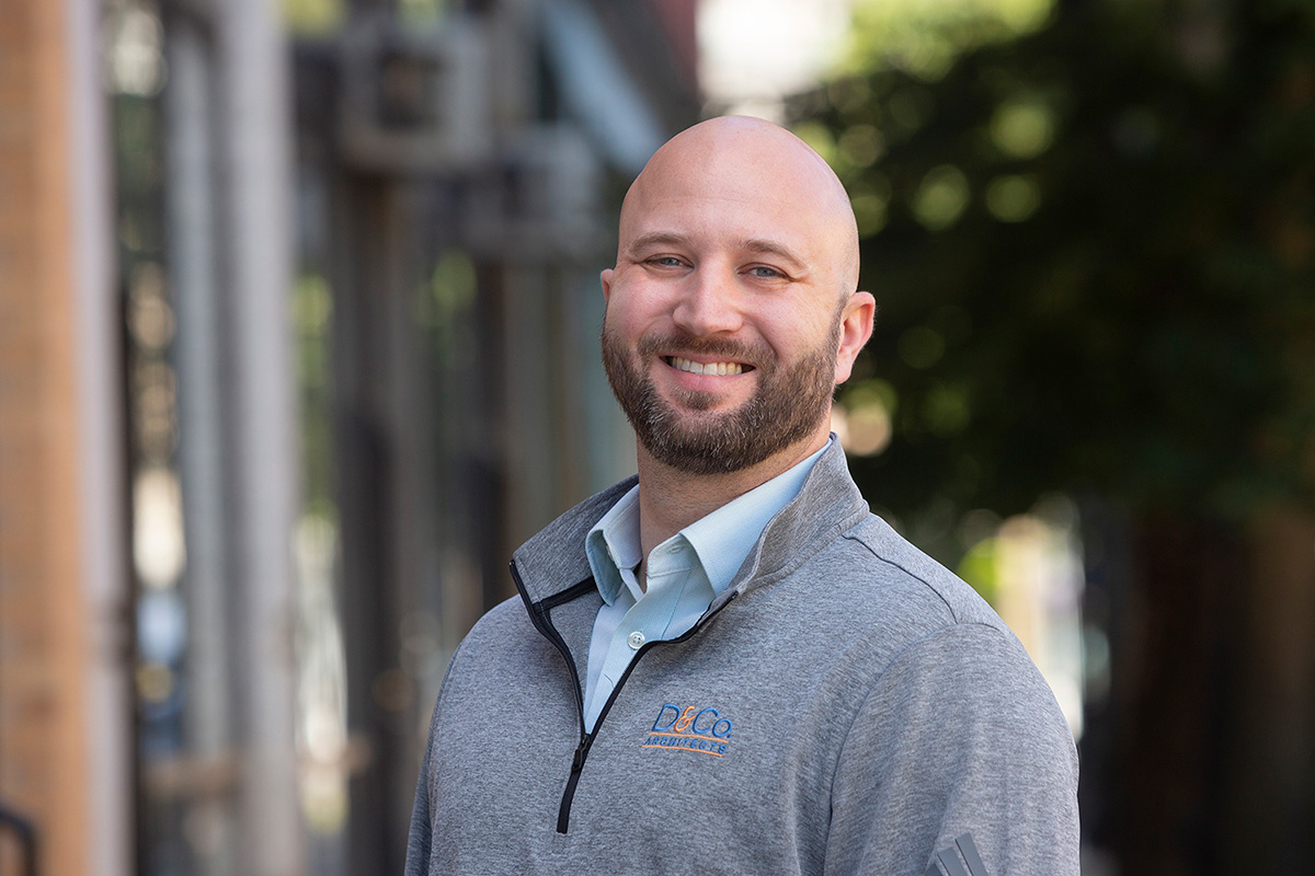 A smiling person with a bald head and beard is wearing a light blue collared shirt under a gray zip-up sweater with a logo, standing outdoors with blurred buildings and greenery in the background.