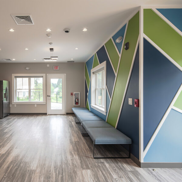Modern kitchen and gathering area in a community building with geometric blue and green accent wall, gray bench seating, and wood flooring.