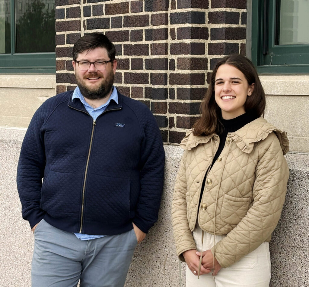 Two people standing in front of a brick and stone building, the man on the left and woman on the right.