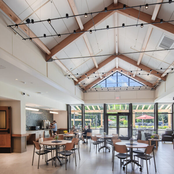 Modern dining area with round tables, wooden beams, large windows, and view of outdoor seating with umbrellas.