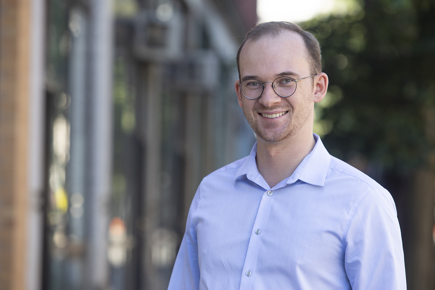 A man with short hair and glasses in a light blue button-up shirt stands outdoors in an urban setting with buildings and trees.