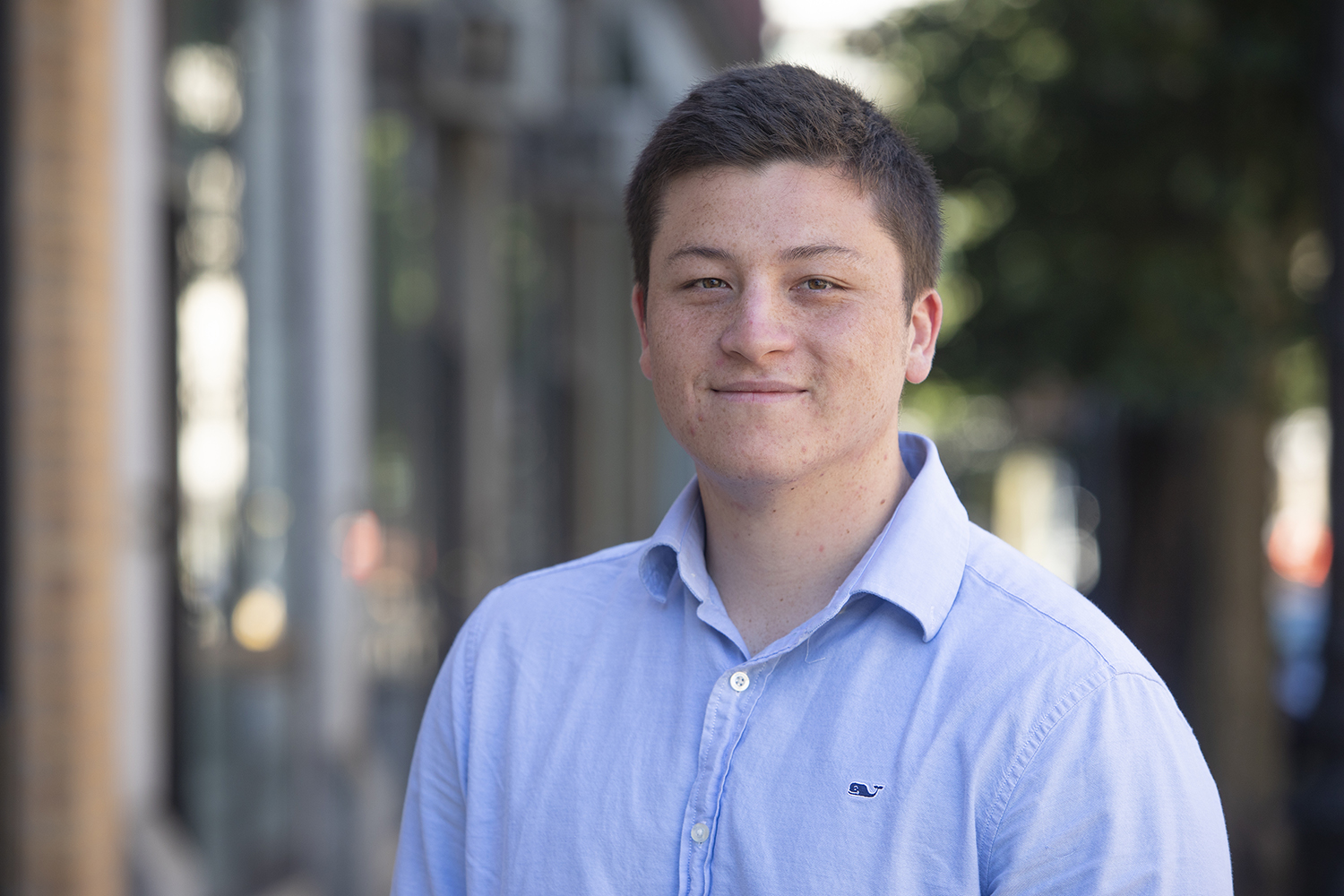 A man in a light blue button-up shirt with a logo stands outdoors in an urban setting with blurred buildings and greenery.