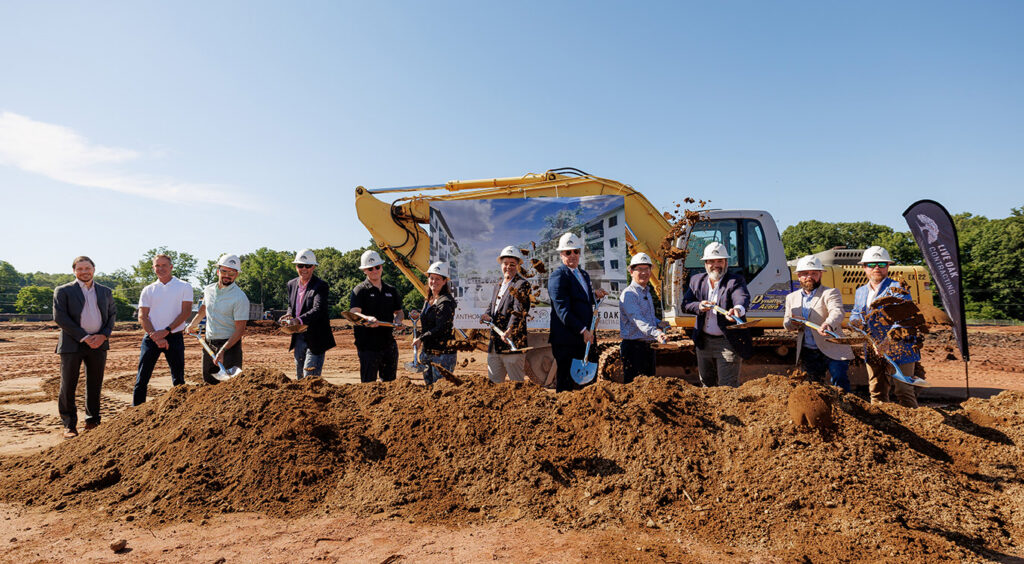 a row of people standing in front of construction equipment and behind a pile of dirt . They are holding shovels and tossing dirt in the air.