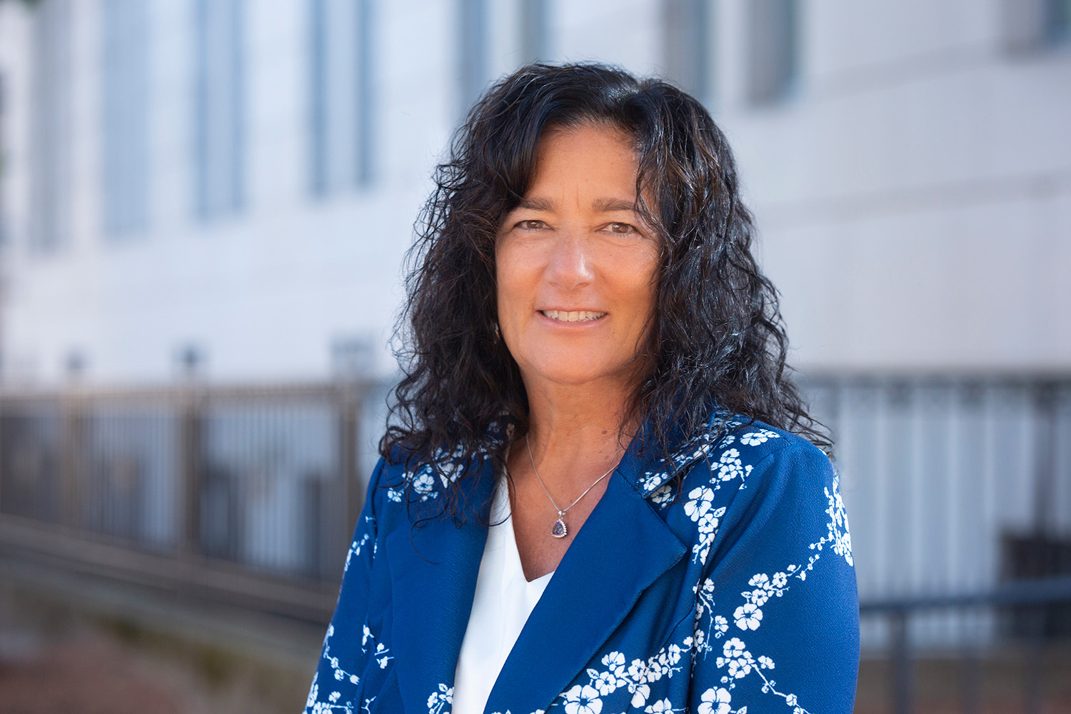 A person with curly dark hair wearing a blue floral blazer and white top is smiling outdoors in front of a building.