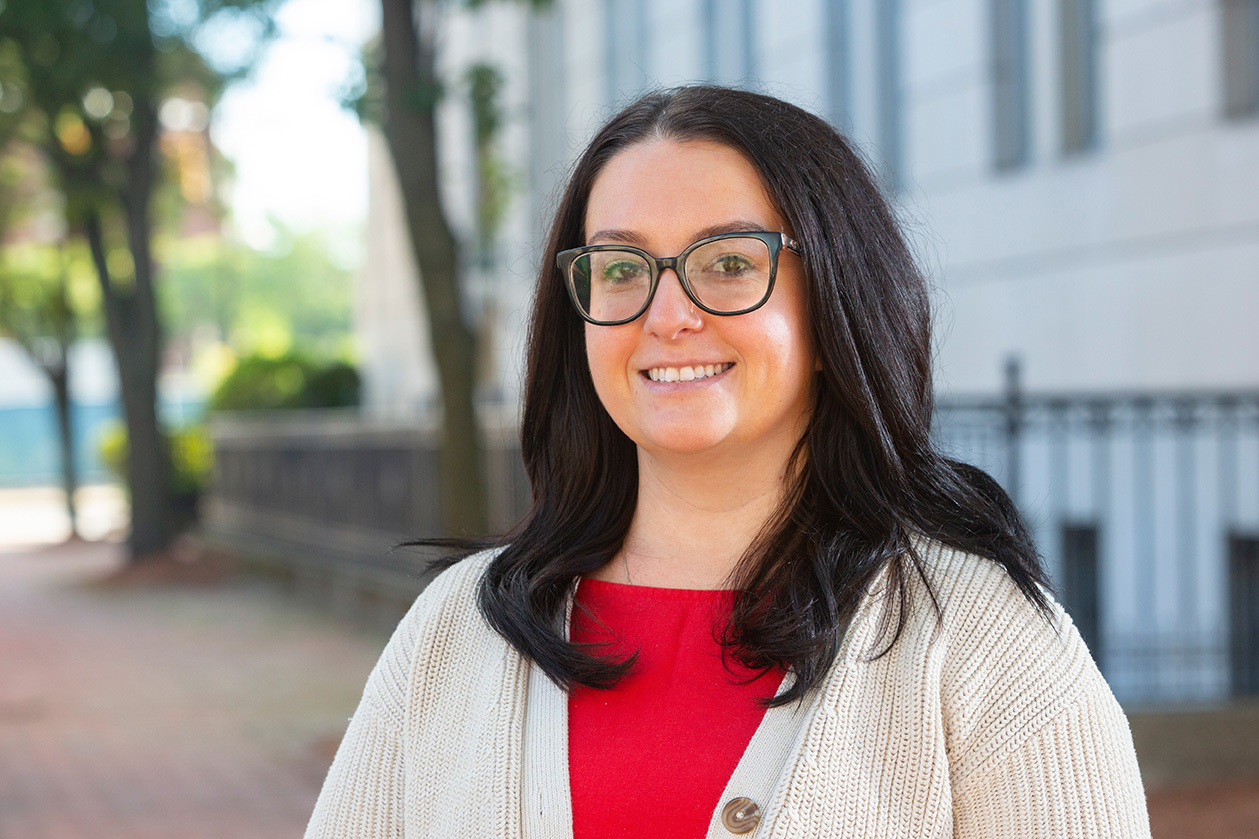 A person with long dark hair and glasses is smiling while wearing a red top and a beige cardigan outdoors near a building and trees.