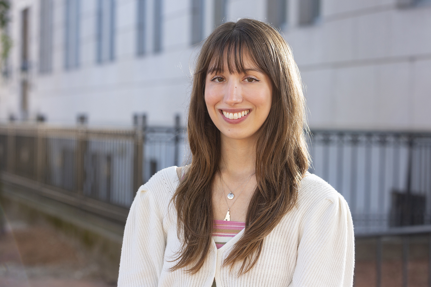 A person with long straight hair and bangs is smiling while wearing a white textured sweater and standing outdoors near a building with a metal fence.