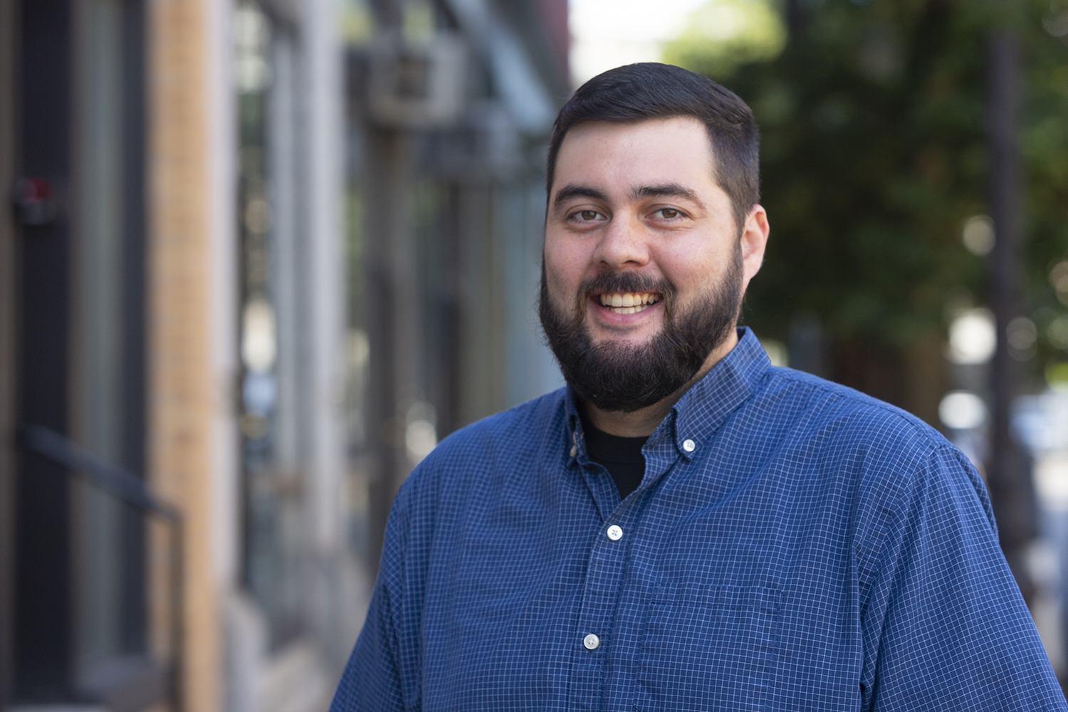 A person with short hair and a beard is smiling and wearing a blue button-up shirt while standing outdoors near buildings and trees.