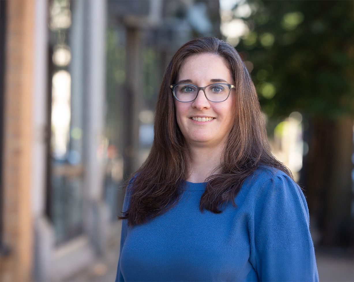 A person with long hair and glasses is smiling while wearing a blue sweater outdoors with blurred buildings and trees in the background.