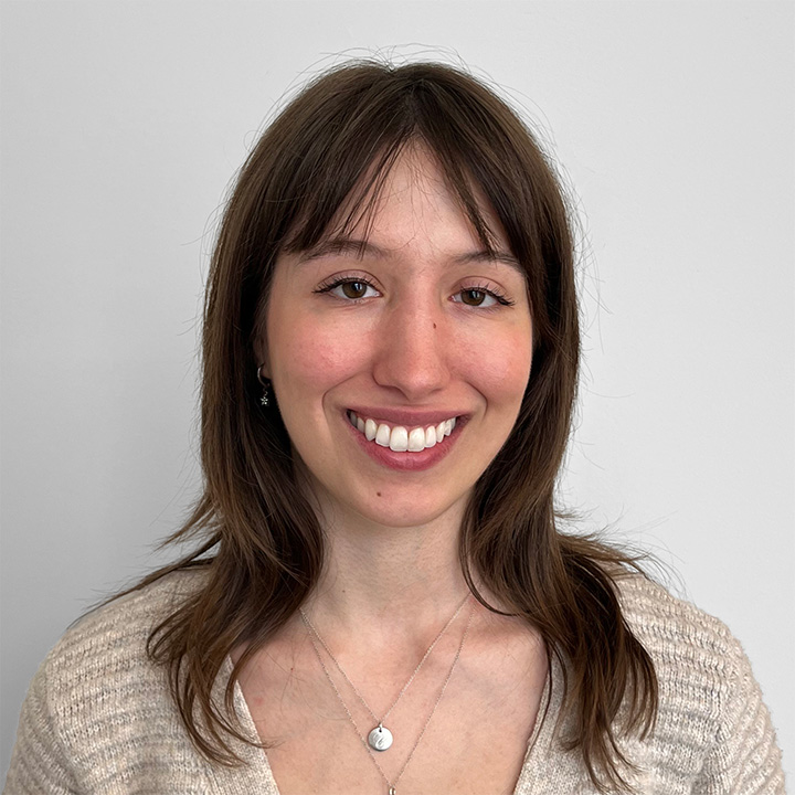 A person with shoulder-length brown hair and bangs is smiling and wearing a beige knit sweater and two silver necklaces against a plain light gray background.
