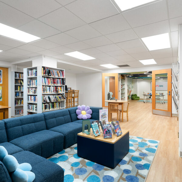 A modern library lounge area features a large blue sectional sofa with flower-shaped pillows, a patterned rug, a small table with books, and bookshelves lining the walls under bright ceiling lights.