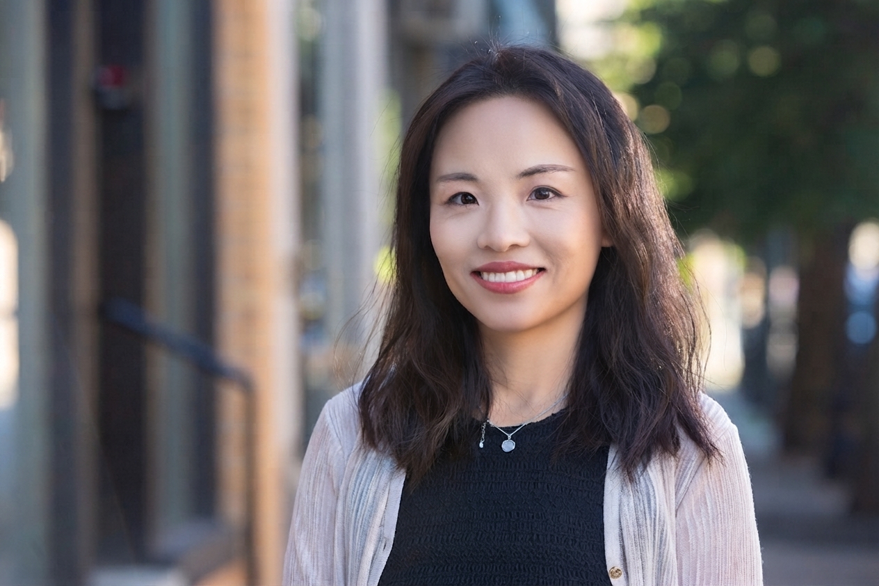 A person with shoulder-length wavy hair wearing a black top and light cardigan is smiling outdoors on a city street.